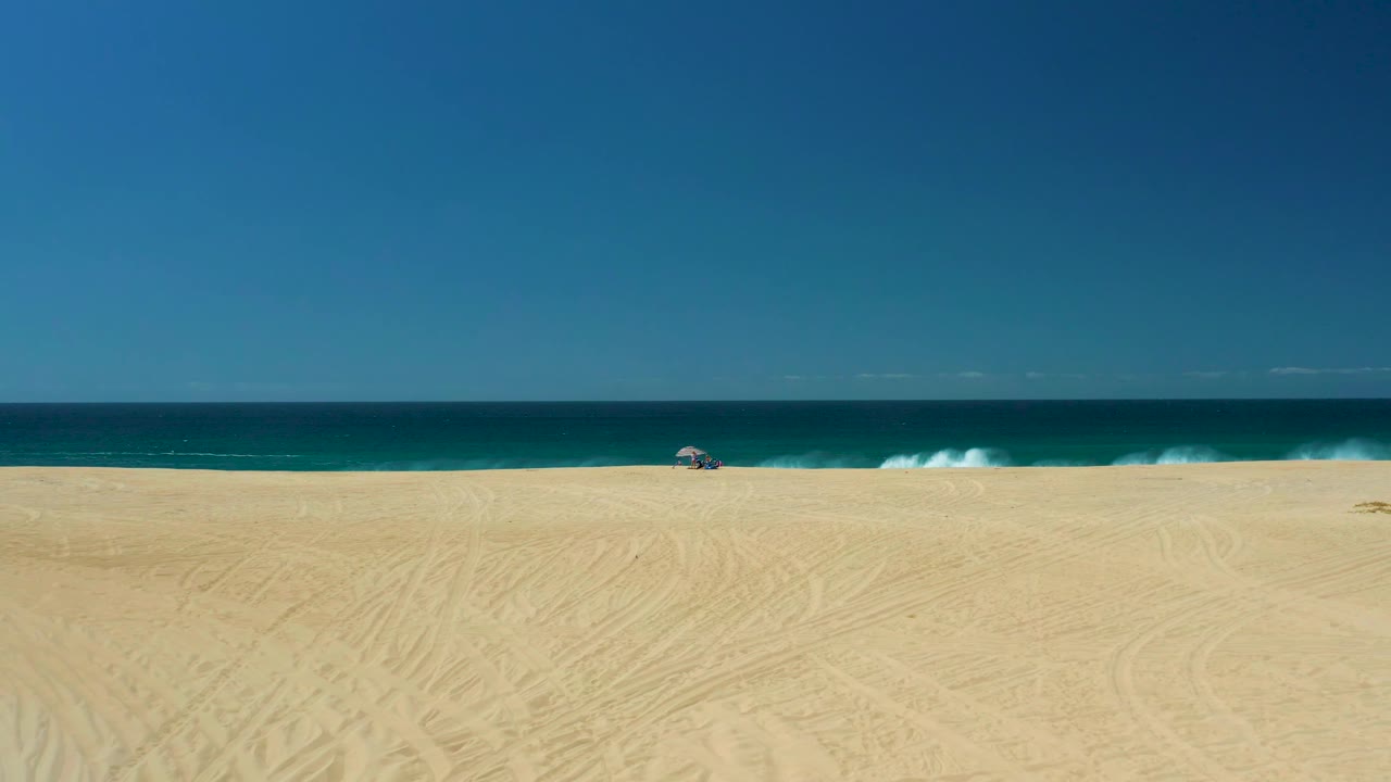 La Laguna Beach shoreline in Los Cabos, Mexico, showing pale sand with track marks, turquoise water near shore, a darker blue horizon, a small umbrella, and no visible people.