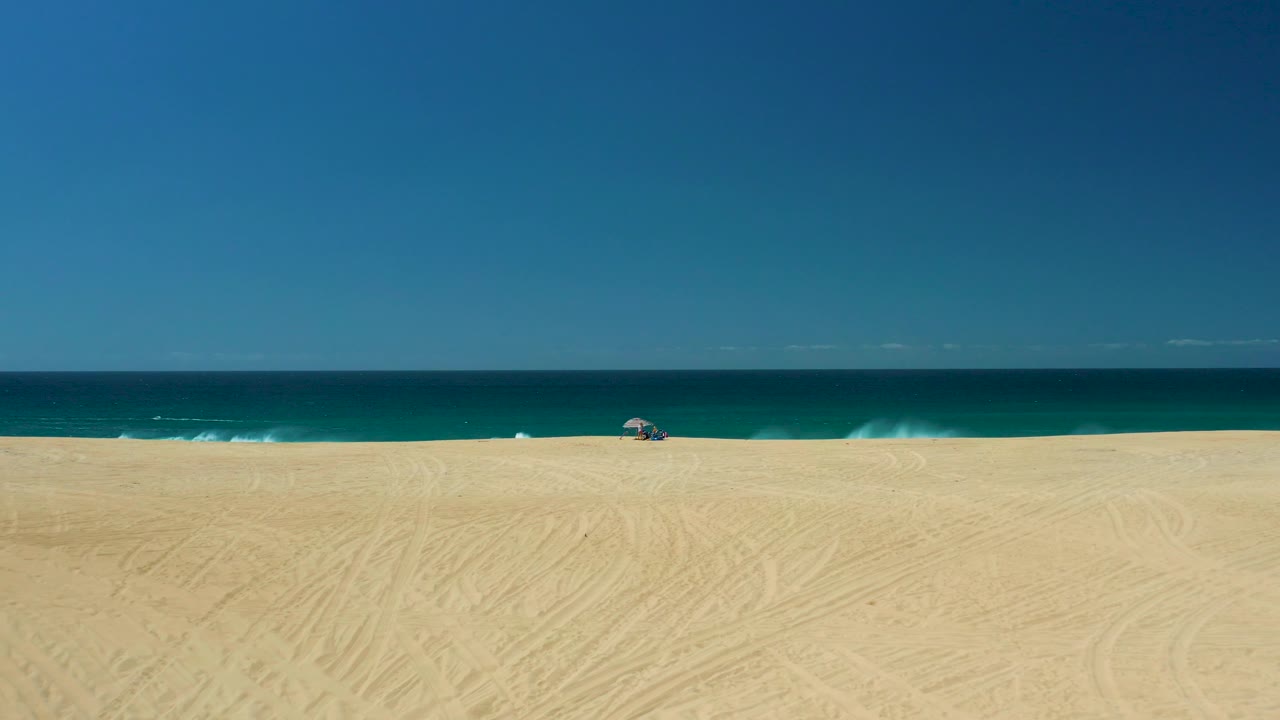 Elevated wide view at La Laguna Beach in Los Cabos, Mexico, with faint sand tracks across the foreground, low white surf along the beach, a distant umbrella, and no visible people.