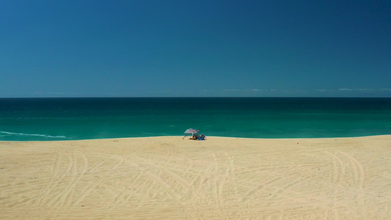 Wide aerial view of La Laguna Beach Los Cabos with pale sand, turquoise ocean, a striped umbrella, small visible beachgoers, and a clear horizon.