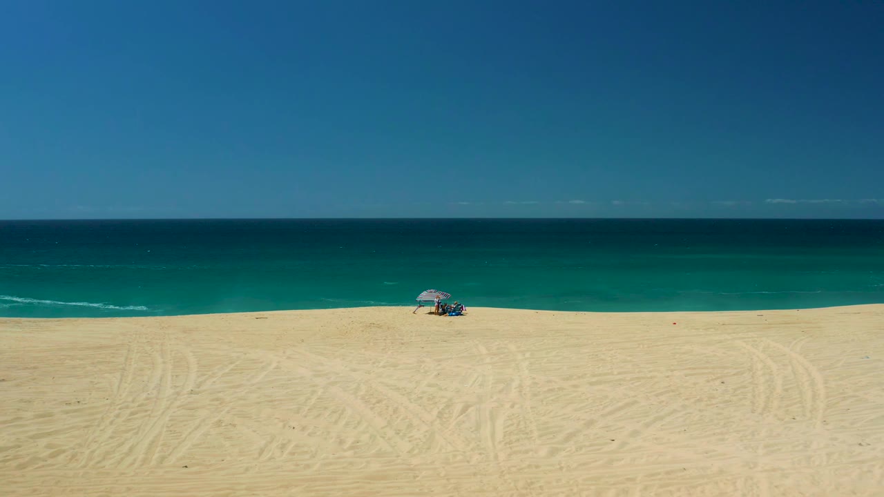 Aerial beach frame with striped umbrella near the shoreline and calm ocean beyond.