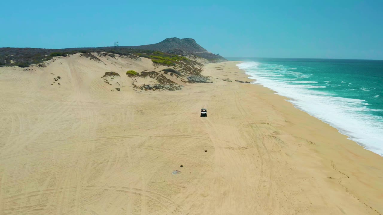 At La Laguna Beach Los Cabos, the car remains centered on the wide sandy beach while white waves break along the right side and dune vegetation lines the left side, with no visible people.