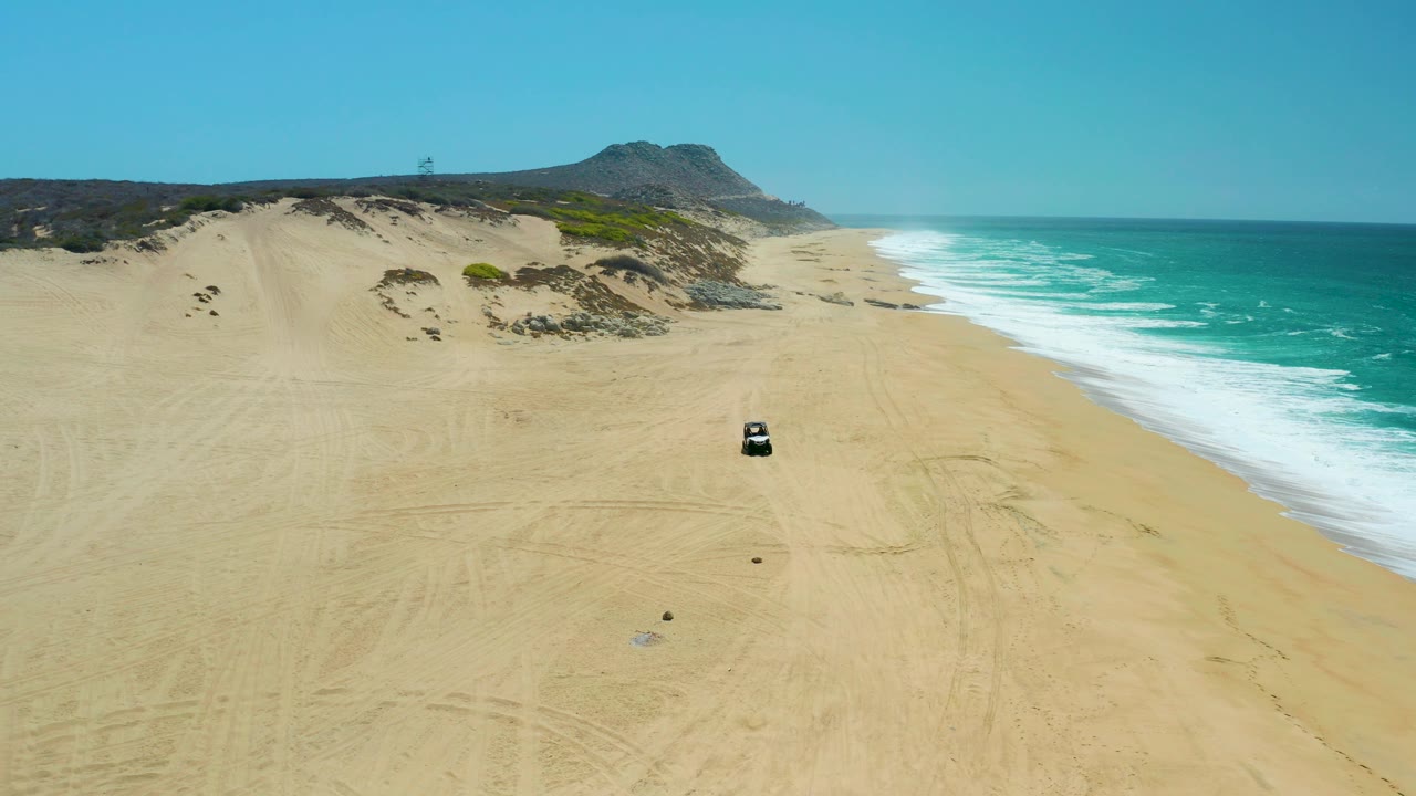 At La Laguna Beach Los Cabos, the vehicle sits slightly closer in the aerial frame, surrounded by pale sand, faint tire marks, bright surf, dune ridges, and no visible people.