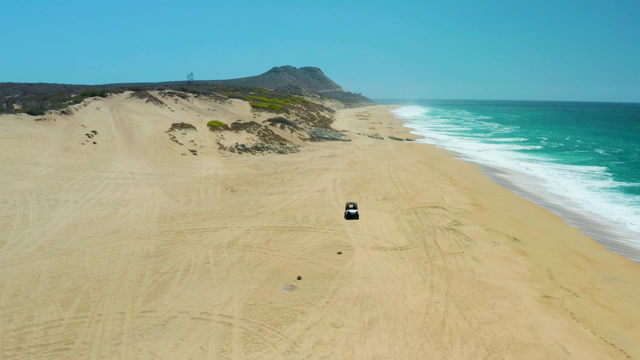At La Laguna Beach Los Cabos, the car is larger in frame as it approaches over tracked sand, with rolling surf on the right, dunes and scrub on the left, and no visible people.