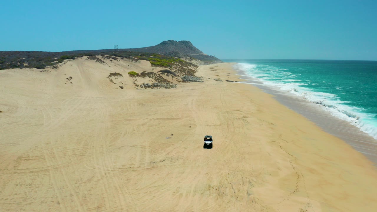 Aerial view of a dark vehicle on a wide sandy beach beside turquoise surf.