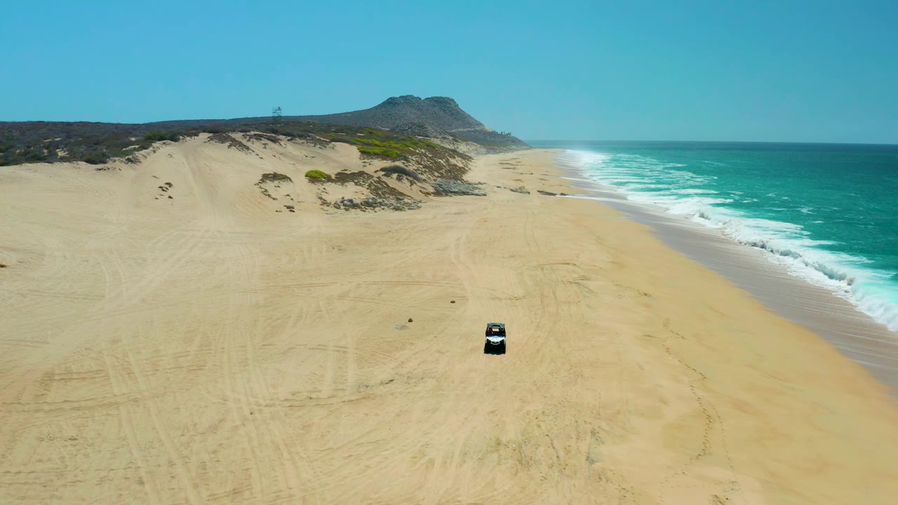 Aerial frame at La Laguna Beach Los Cabos with the dark vehicle slightly farther along the pale beach, white wave foam edging the shore, dunes behind it, and no visible people.