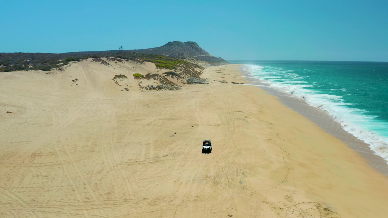 At La Laguna Beach Los Cabos, the aerial view shows the dark vehicle farther down the sandy beach, the surf line closer to frame right, dunes in back, and no visible people.