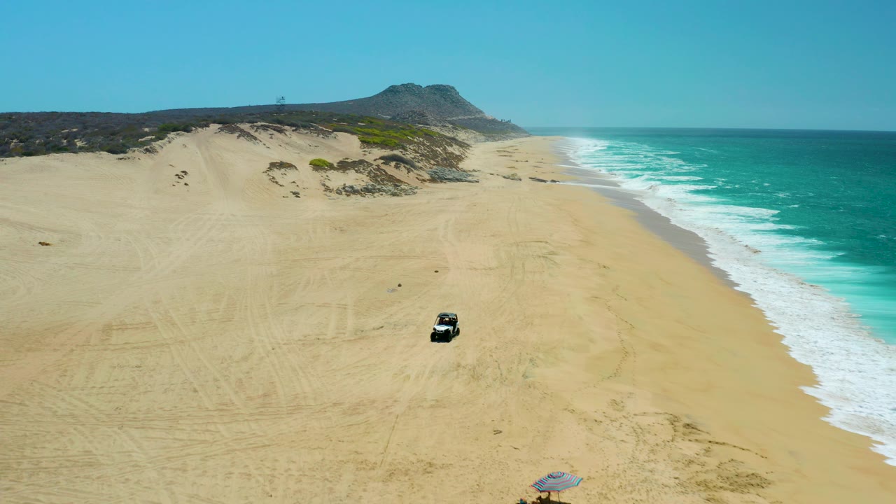 Aerial frame from La Laguna Beach Los Cabos showing the dark vehicle lower in the sand field, bright surf breaking along the right edge, dune slopes beyond, and no visible people.