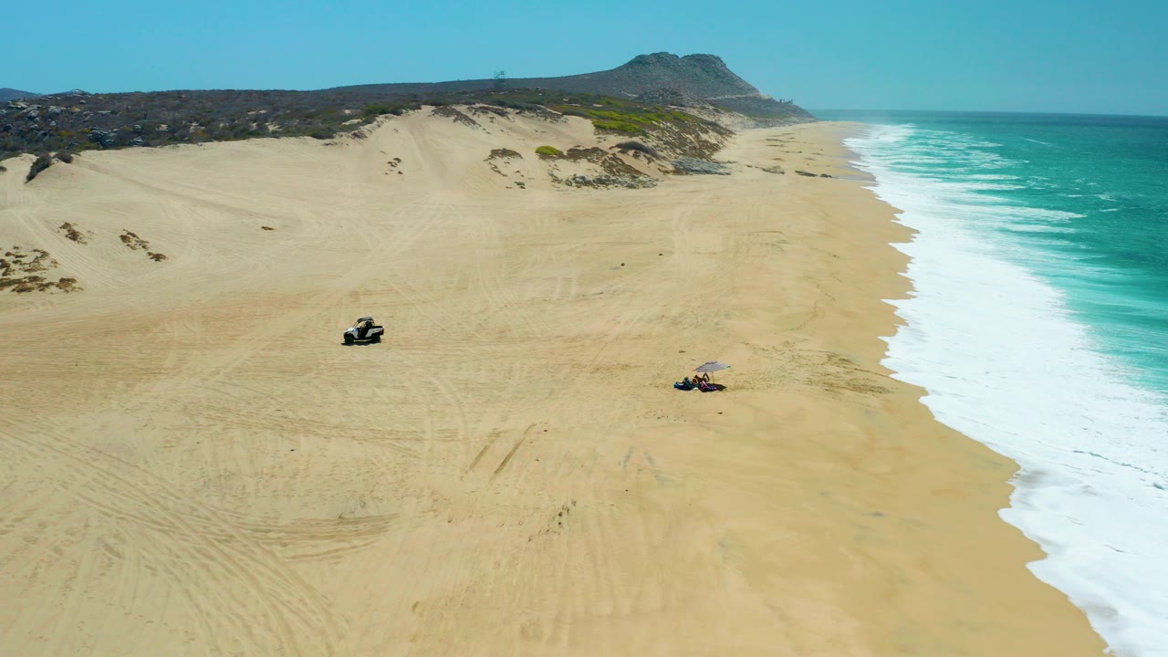 Aerial view at La Laguna Beach Los Cabos showing two ATVs on wide sand between pale dunes and turquoise surf, with distant hills under clear sky and no visible people.
