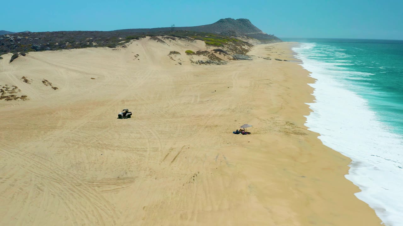 Aerial frame of La Laguna Beach Los Cabos with two ATVs moving across open sand, white waves breaking to the right, dunes and scrub behind them, and no visible people.