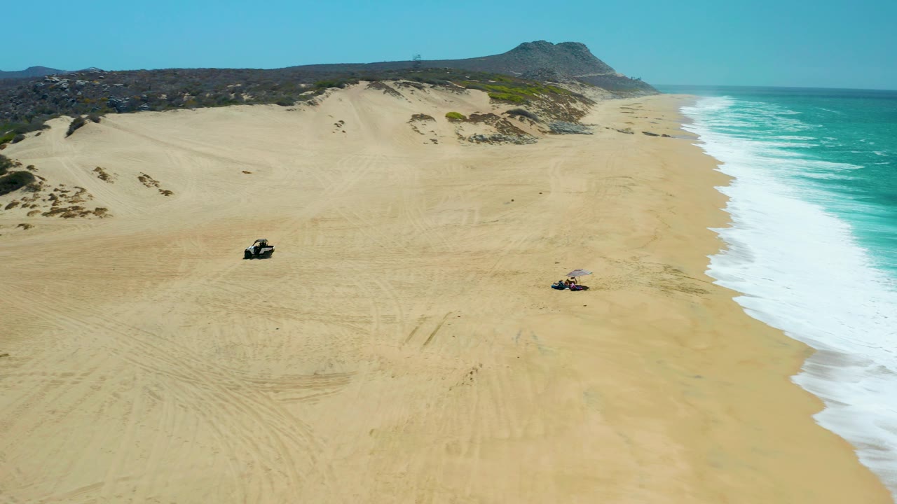 Aerial view over La Laguna Beach Los Cabos as the ATVs sit farther across the sandy expanse, with foamy surf along the right edge, dunes in back, and no visible people.