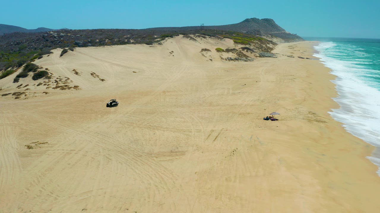 Drone view of two ATVs crossing sand near dunes and surf.