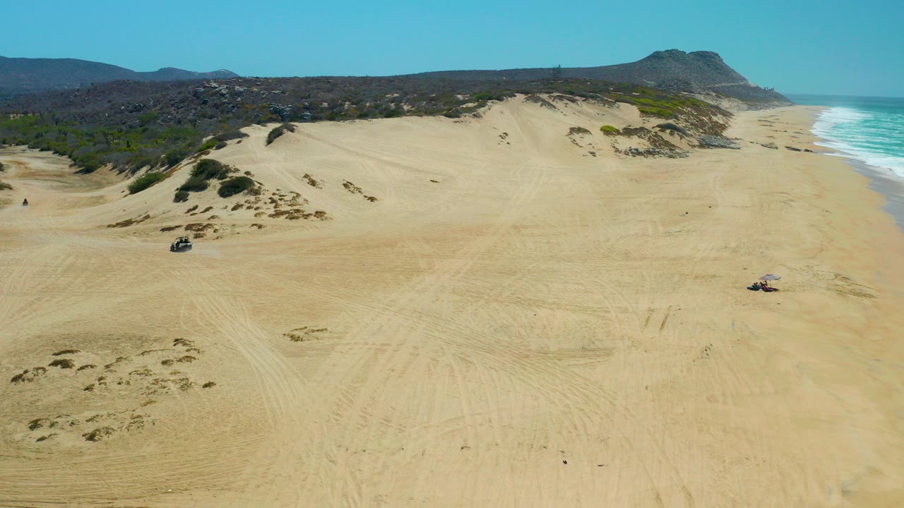 Aerial frame from La Laguna Beach Los Cabos showing broad sand dunes, faint tire tracks, a small 4x4 on the open beach, ocean surf at right, distant hills, and no recognizable people.