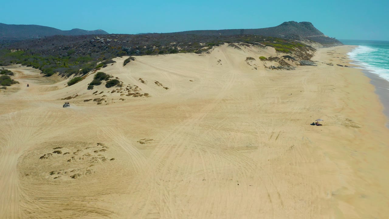 Drone view of broad beach dunes with vehicle tracks and surf along the right side.