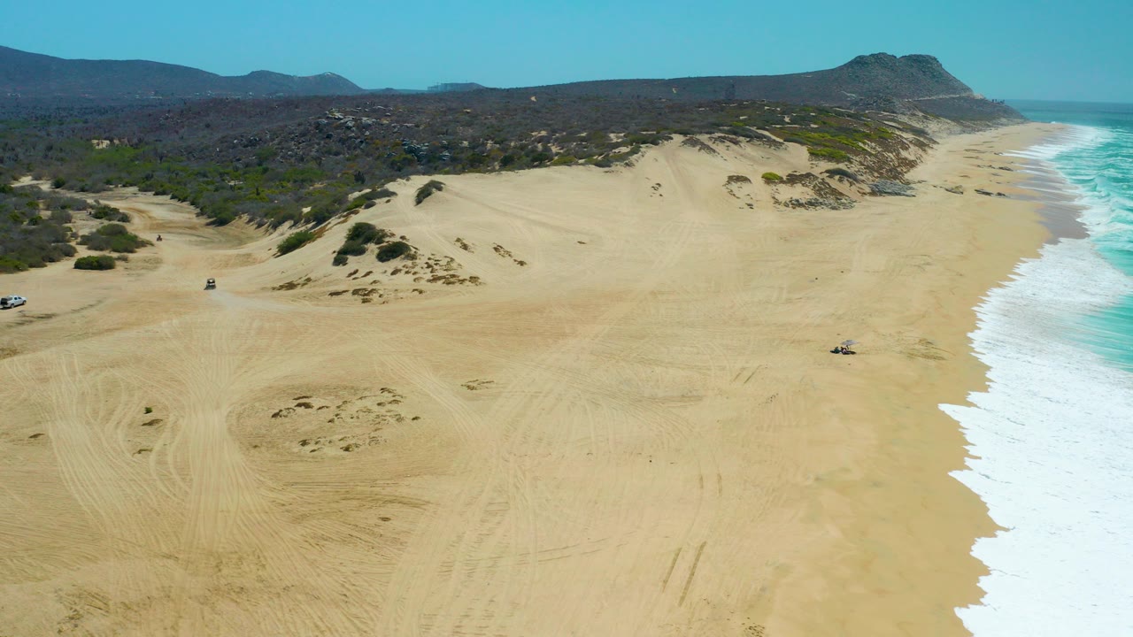 Aerial frame over La Laguna Beach Los Cabos showing broad sand dunes, blue surf along the right edge, distant hills, small vehicles on the beach, and no visible people.