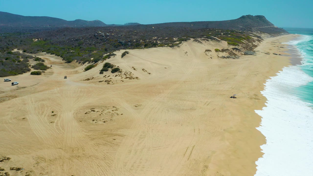 Aerial view of La Laguna Beach Los Cabos with tire-marked sand in the foreground, several vehicles near the left side, turquoise water and white surf to the right, and no visible people.