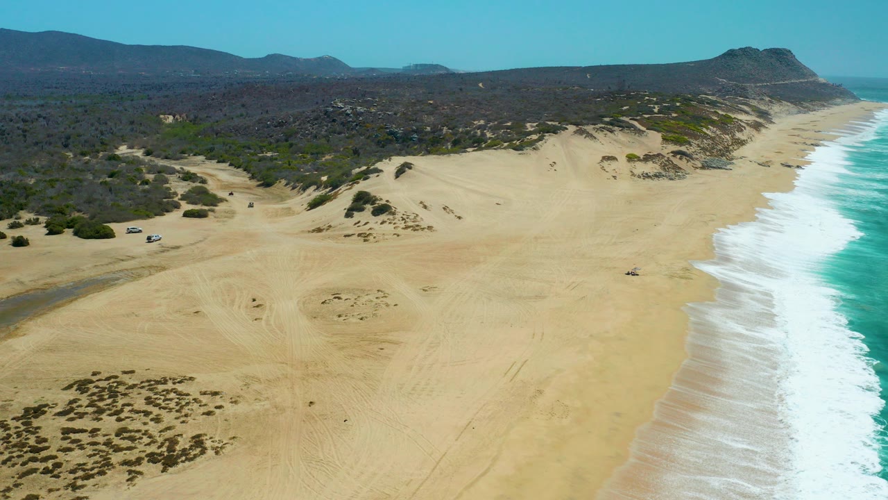 High aerial frame over La Laguna Beach Los Cabos showing pale sand dunes, turquoise surf on the right, inland scrub and hills, and no visible people.