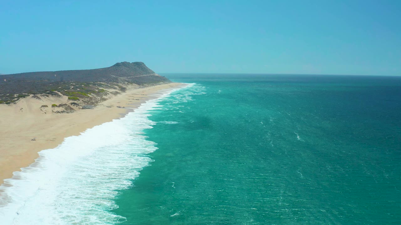 At 0:01, La Laguna Beach Los Cabos remains framed from above with the surf line slightly farther down the beach, deep turquoise water to the right, sand to the left, and no visible people.