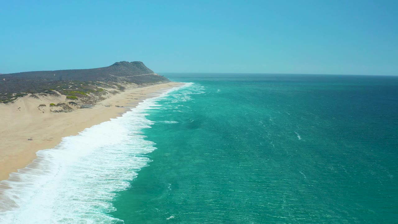 At 0:02, the La Laguna Beach Los Cabos shoreline shows a broad white foam strip, pale sand, textured turquoise water, distant low terrain, and no visible people.