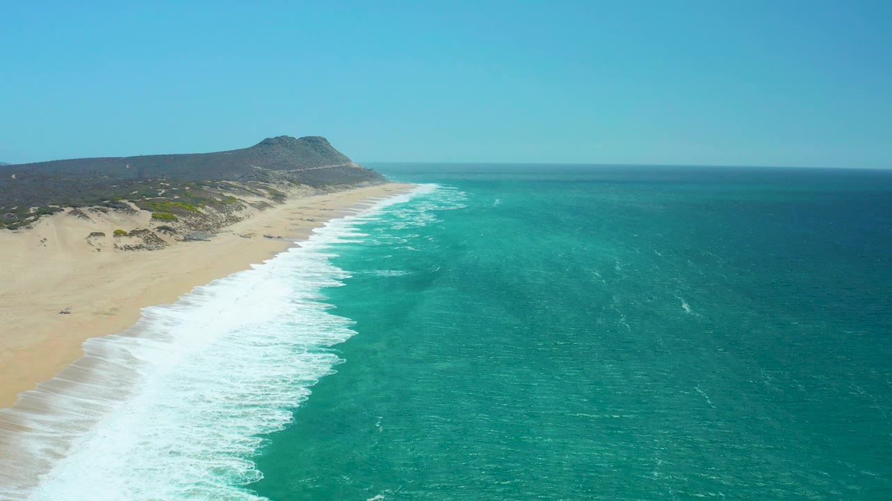 At 0:03, the aerial view over La Laguna Beach Los Cabos shows the beach stretching into the distance, rolling surf along the left edge of the water, open sea to the right, and no visible people.
