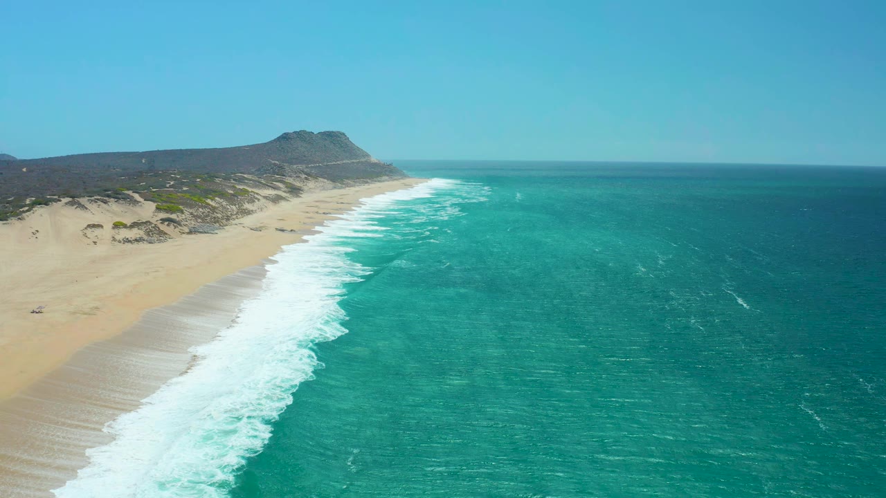 At La Laguna Beach Los Cabos, white foam has advanced farther along the beach while the turquoise ocean fills the right side and the dune-backed coast remains visible, with no visible people.