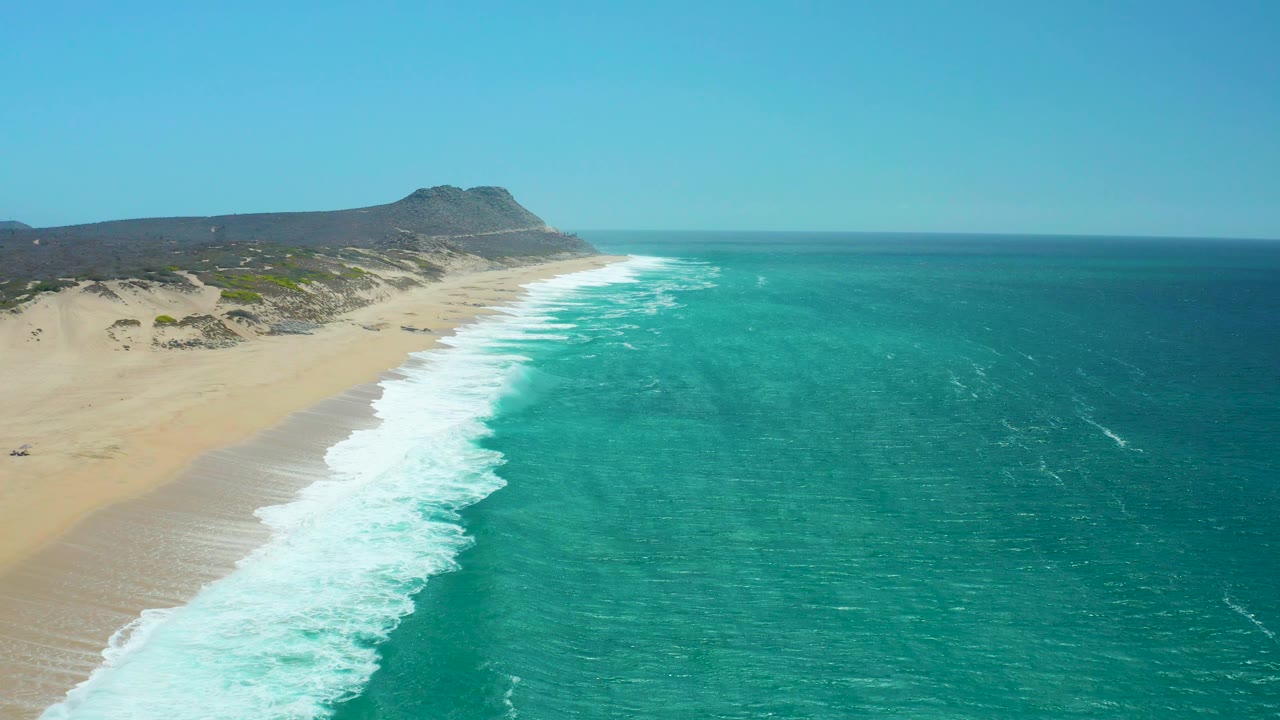 At La Laguna Beach Los Cabos, foamy waves push up the left side of the sandy shore while low hills and scrub-covered dunes sit behind the beach, with no visible people.