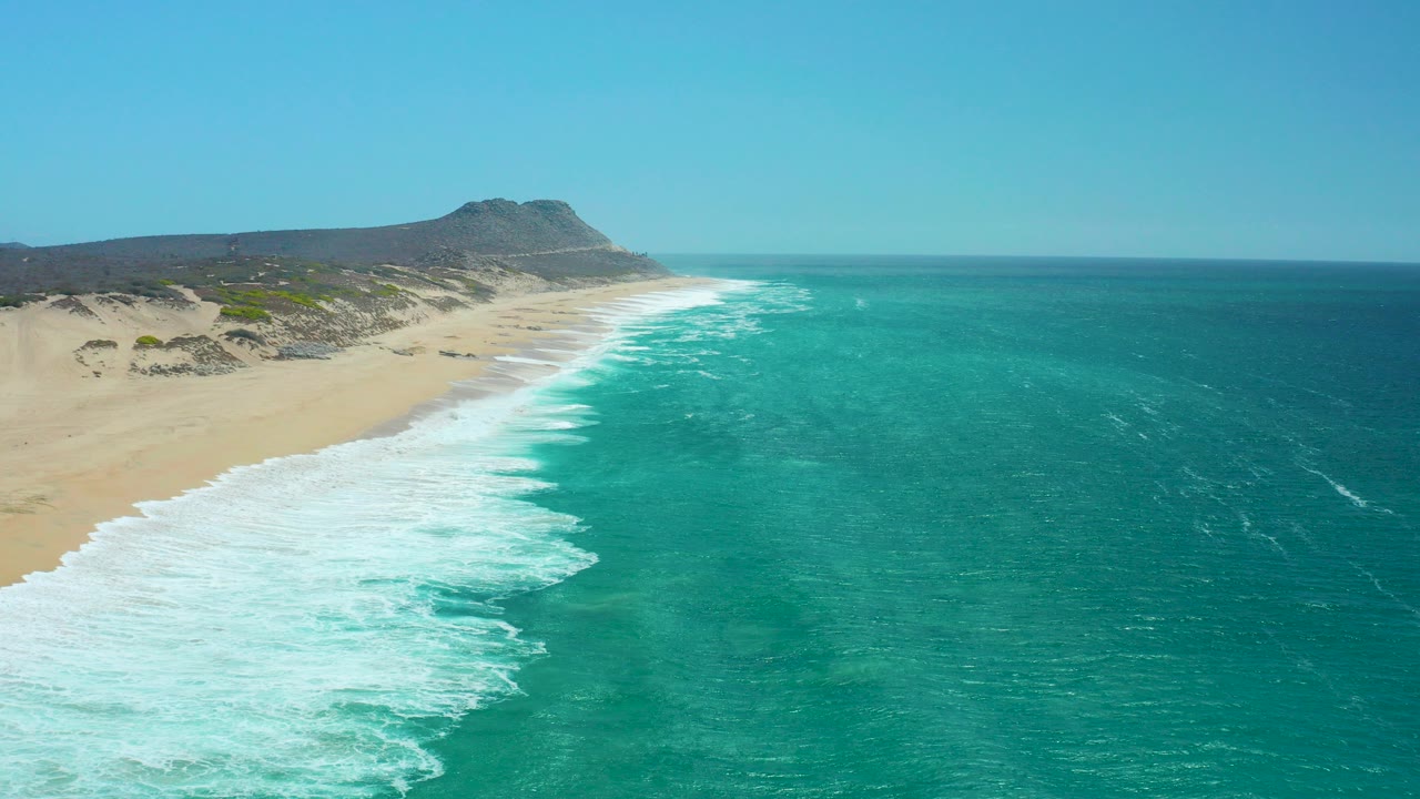 Aerial view at La Laguna Beach Los Cabos showing turquoise ocean, white surf, sandy shore, dunes, a distant hill, and no visible people.