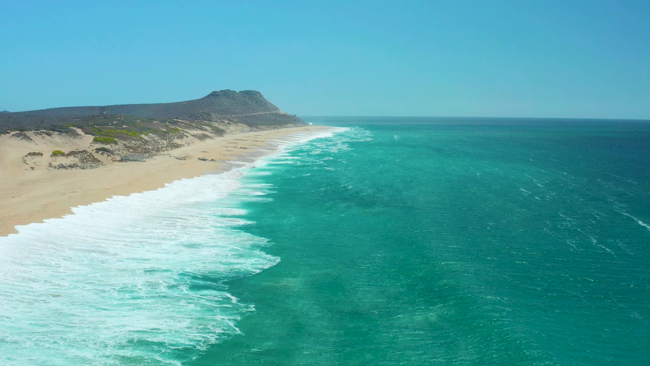 La Laguna Beach Los Cabos seen from above with the shore running from foreground left toward a dark hill, white breakers along the sand, clear sky, and no visible people.