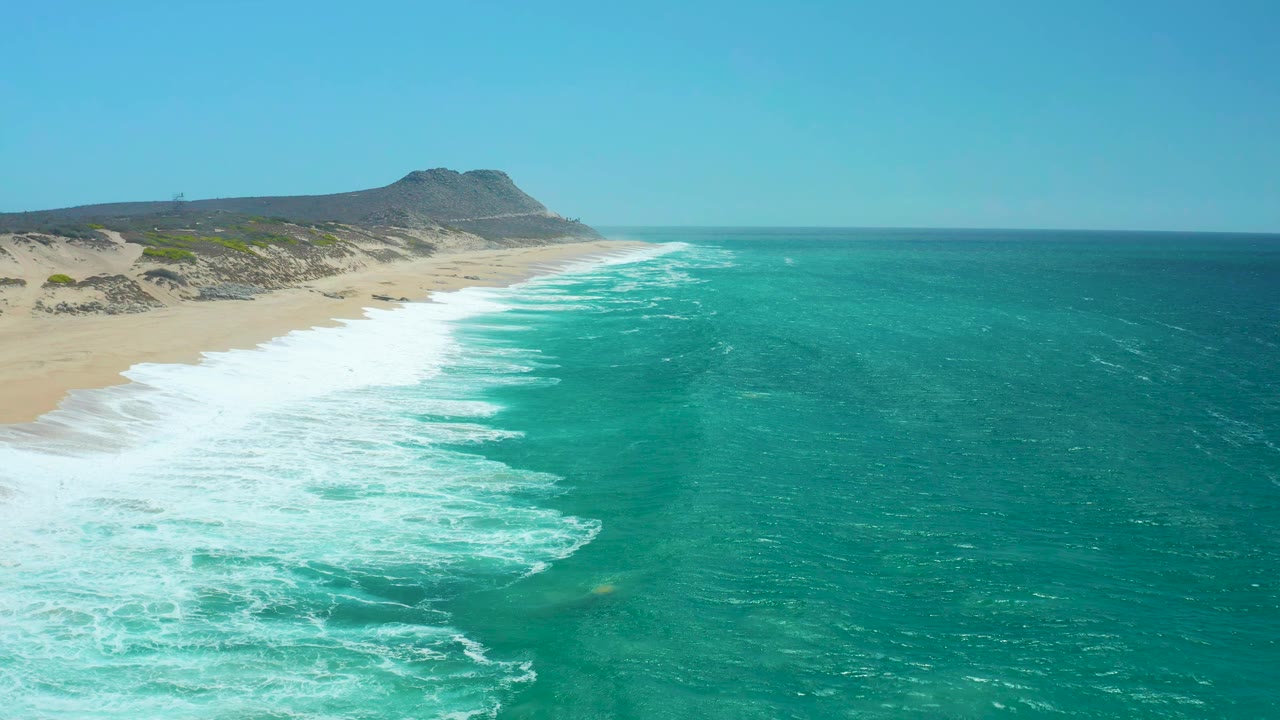 Wide aerial coastline with white foam spreading across turquoise water and sand.