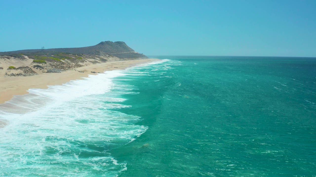 Aerial view of La Laguna Beach Los Cabos with surf curling toward the beach, a broad turquoise water field to the right, and no visible people.