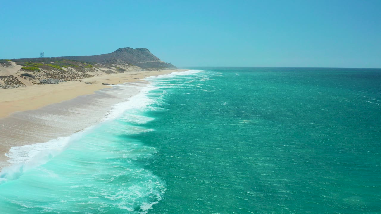 Aerial view of turquoise waves breaking along a sandy beach below a clear sky.