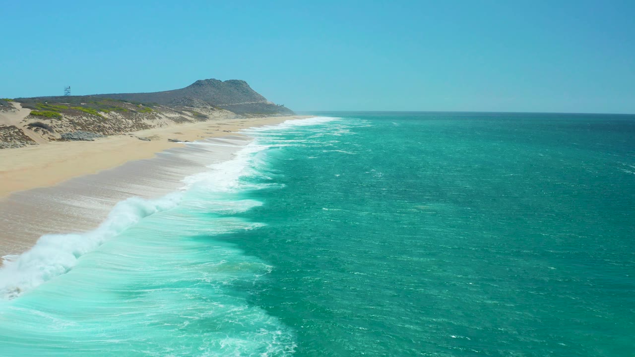 At 0:02, the surf thickens near the lower left edge of the La Laguna Beach Los Cabos shoreline, with clear sky above the dunes and no visible people on the sand.