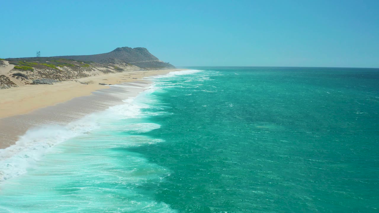 At 0:03, the aerial angle keeps the beach on the left and the blue-green ocean on the right as white water pushes up the La Laguna Beach Los Cabos shore with no visible people.