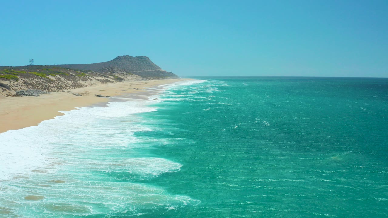 Opening frame of La Laguna Beach in Los Cabos, Mexico, with turquoise water, broad white foam, sandy shore, low dunes, distant hill, and no visible people.
