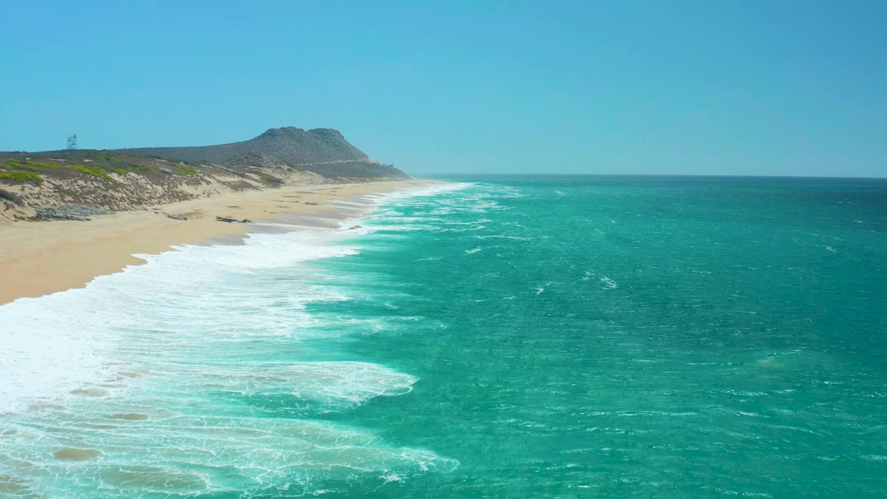 A second moment at La Laguna Beach shows white breakers spreading farther across the sand, turquoise ocean on the right, dunes and hill in the background, and no visible people.