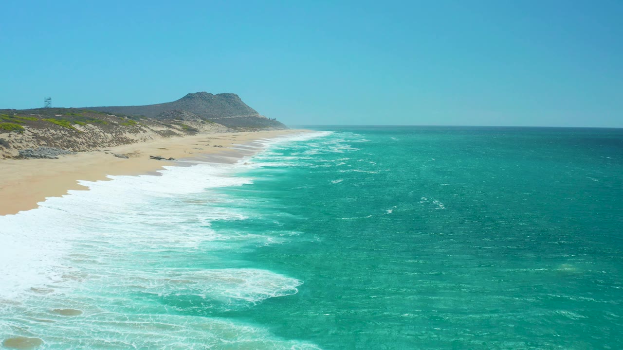 The La Laguna Beach shoreline continues under clear sky, with a bright band of surf curling toward the sand, open turquoise water to the right, and no visible people.