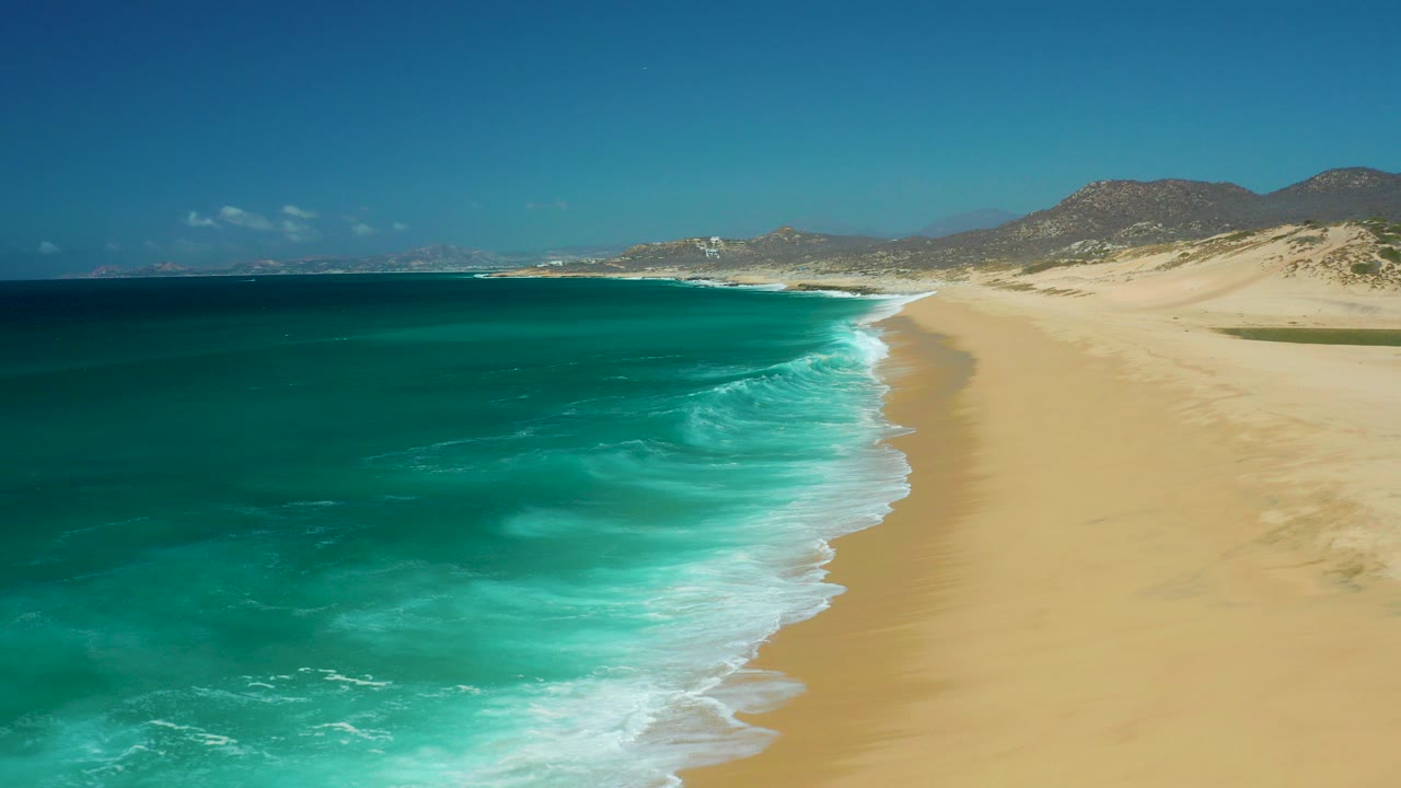Aerial view of turquoise surf meeting a sandy beach with hills in the distance.