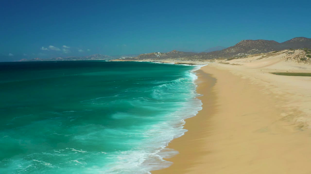 At 0:01, La Laguna Beach Los Cabos appears from above with deeper blue water offshore, bright turquoise shallows, a pale sand beach, distant hills, and no visible people.
