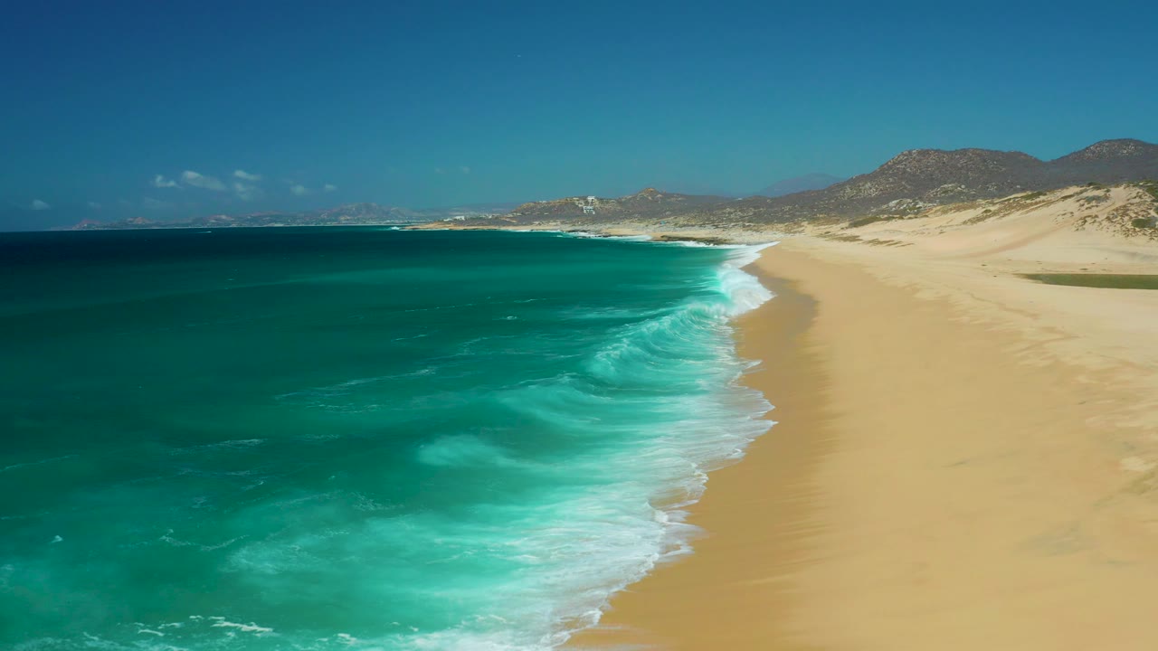 At La Laguna Beach Los Cabos, a foamy wave band reaches farther along the sand while the turquoise ocean fills the left side and low hills sit below a clear sky, with no visible people.