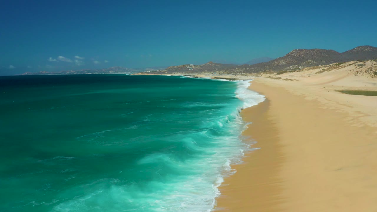 At 0:03, the aerial view looks along La Laguna Beach Los Cabos with dark blue water offshore, turquoise surf near the beach, broad sand to the right, hills beyond, and no visible people.
