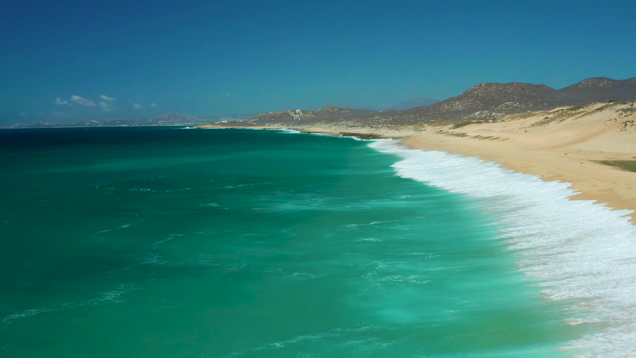 La Laguna Beach Los Cabos shoreline seen from above, with a broad band of white foam pushing toward the sandy beach and low brown hills beyond, with no visible people.