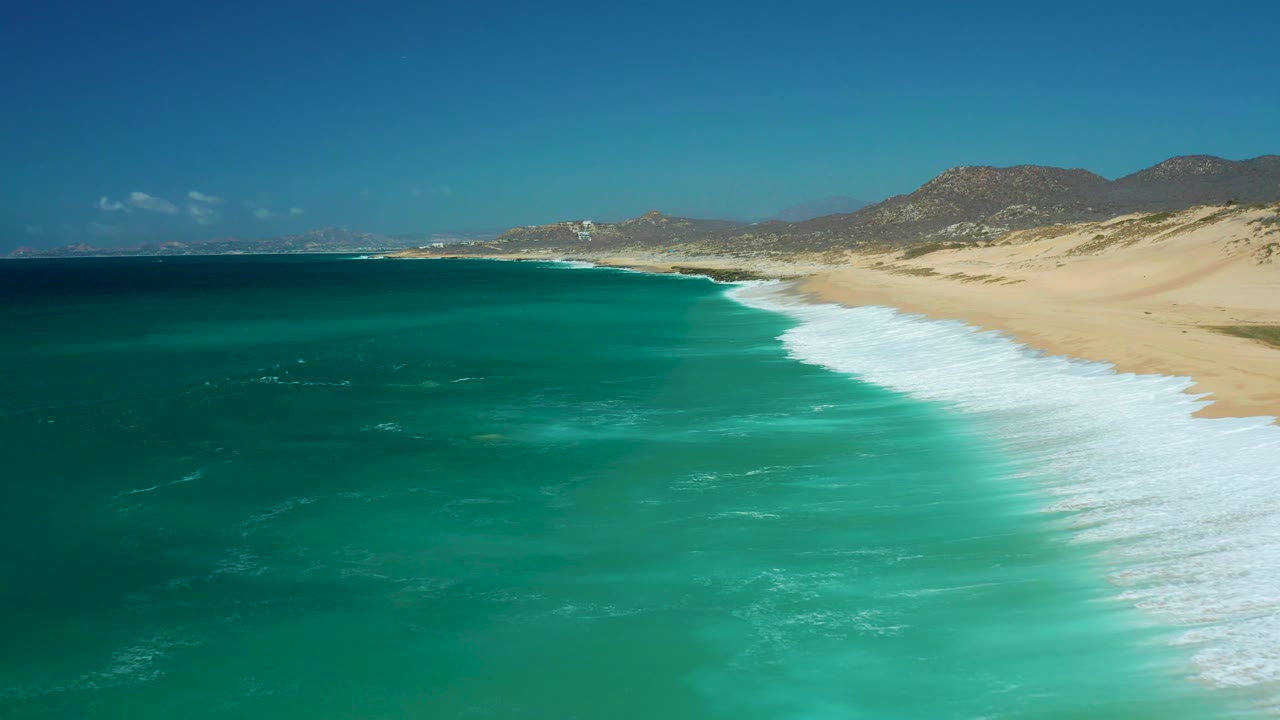 Aerial frame of La Laguna Beach Los Cabos with darker offshore water at left, bright turquoise shallows near the surf, a long sandy beach at right, and no visible people.