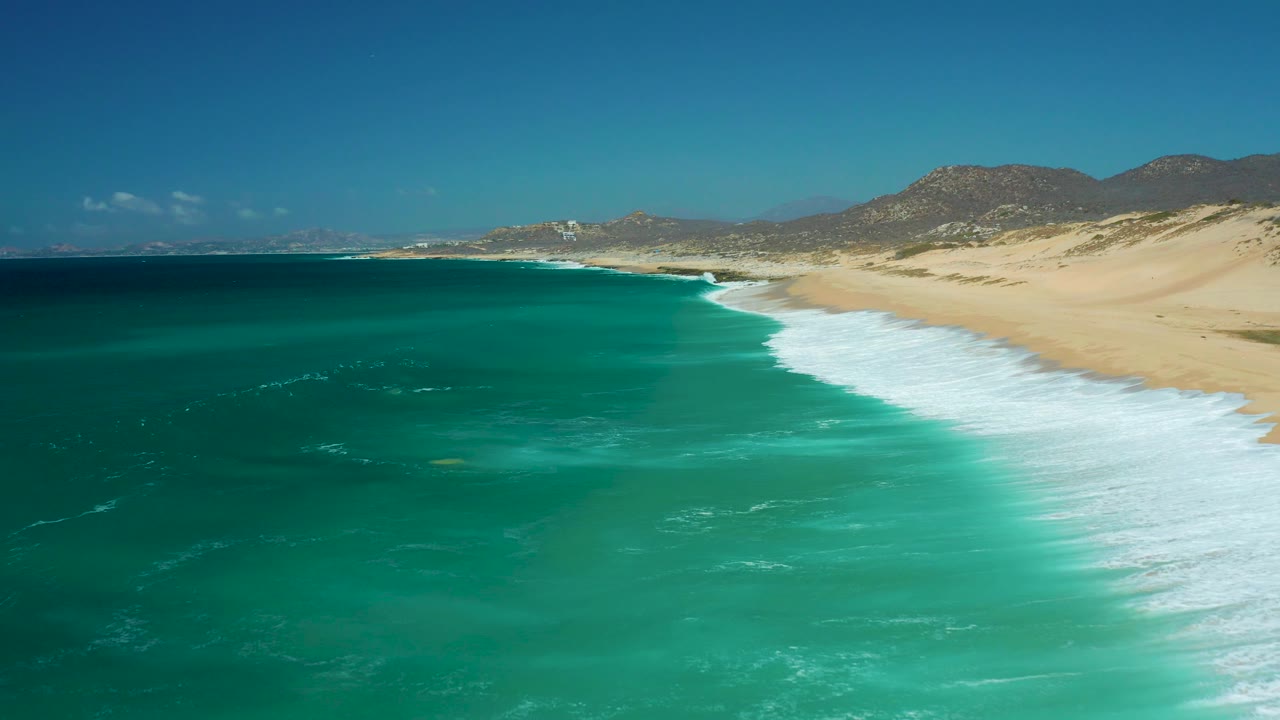 La Laguna Beach Los Cabos coastline from a drone perspective, with open turquoise water on the left, a clean white surf edge, pale sand on the right, and no visible people.