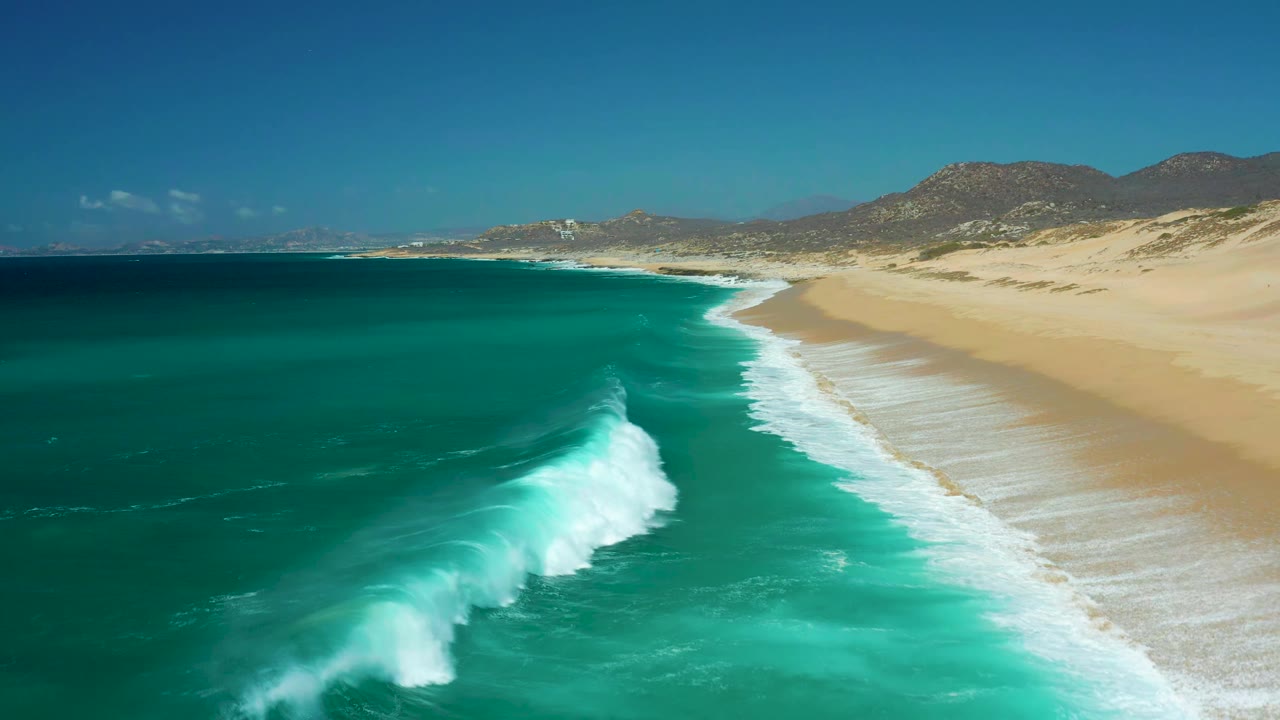 Aerial shoreline view at La Laguna Beach Los Cabos, Mexico, as a white surf line rolls diagonally toward the tan beach, with open teal water, dunes, hills, and no visible people.