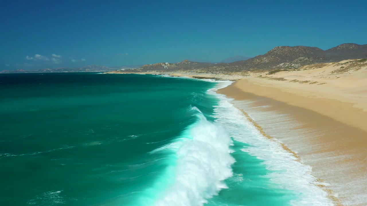 Aerial frame from La Laguna Beach Los Cabos, Mexico, with a larger white wave crest rising near the center, turquoise water around it, sandy beach to the right, distant hills, and no visible people.