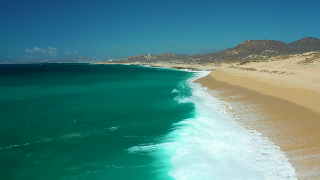 Aerial view over La Laguna Beach Los Cabos, Mexico, with foamy surf spreading down the right side of the frame, darker open water on the left, tan sand, distant hills, and no visible people.