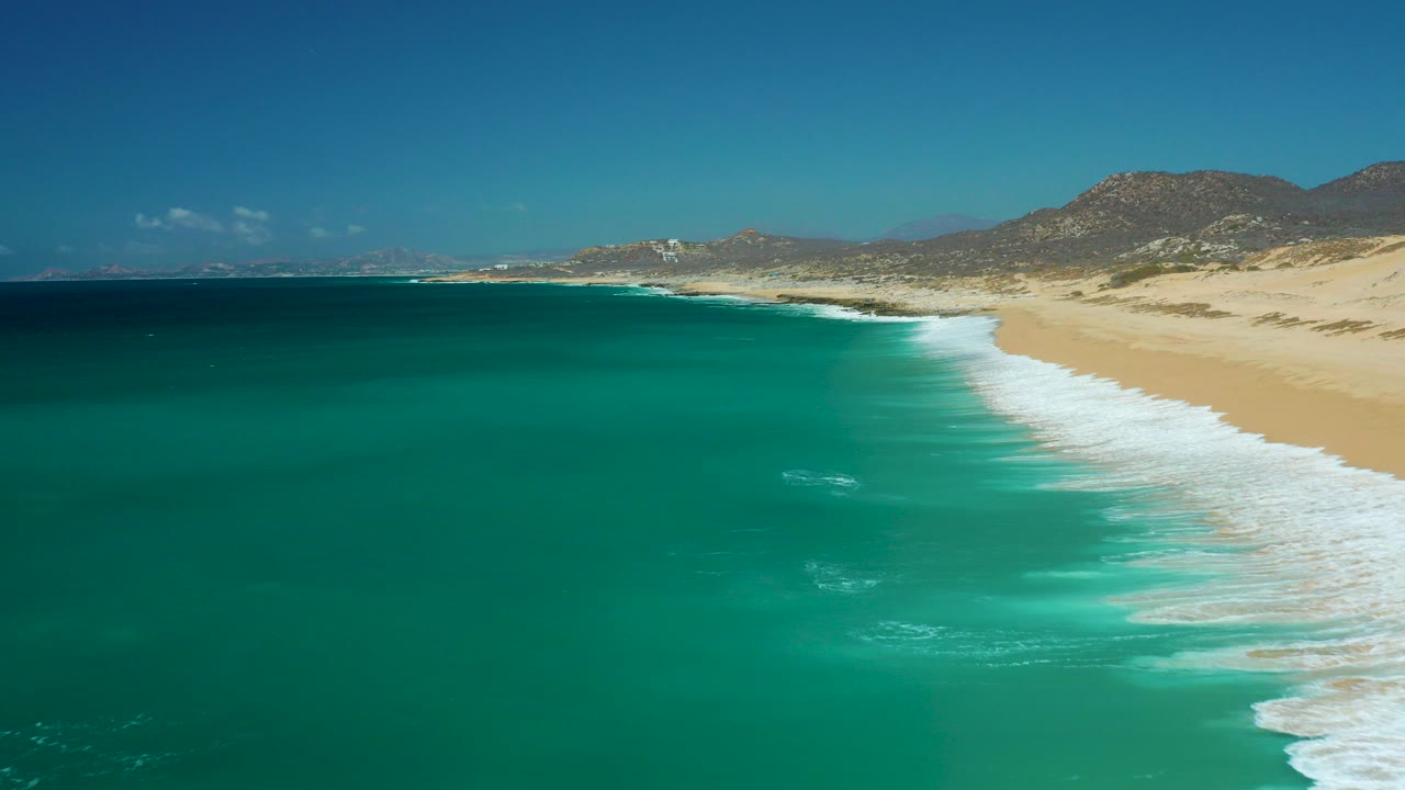 At La Laguna Beach Los Cabos, turquoise water fills the foreground while white surf reaches the sandy shore and mountains line the background, with no visible people.