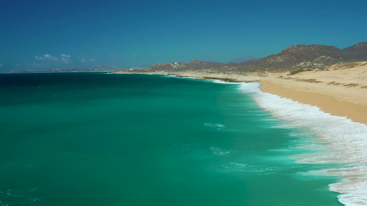 At La Laguna Beach Los Cabos, the surf line sits farther down the right edge as turquoise water spreads across the frame, with low mountains and clear sky behind, and no visible people.
