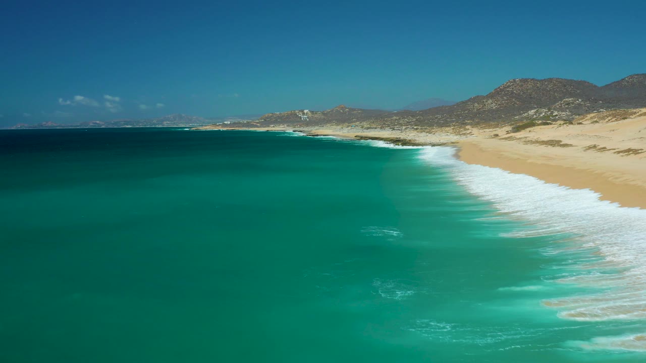 At La Laguna Beach Los Cabos, white water gathers along the right-hand beach while deeper teal ocean occupies the left and mountains remain visible under a clear sky, with no visible people.