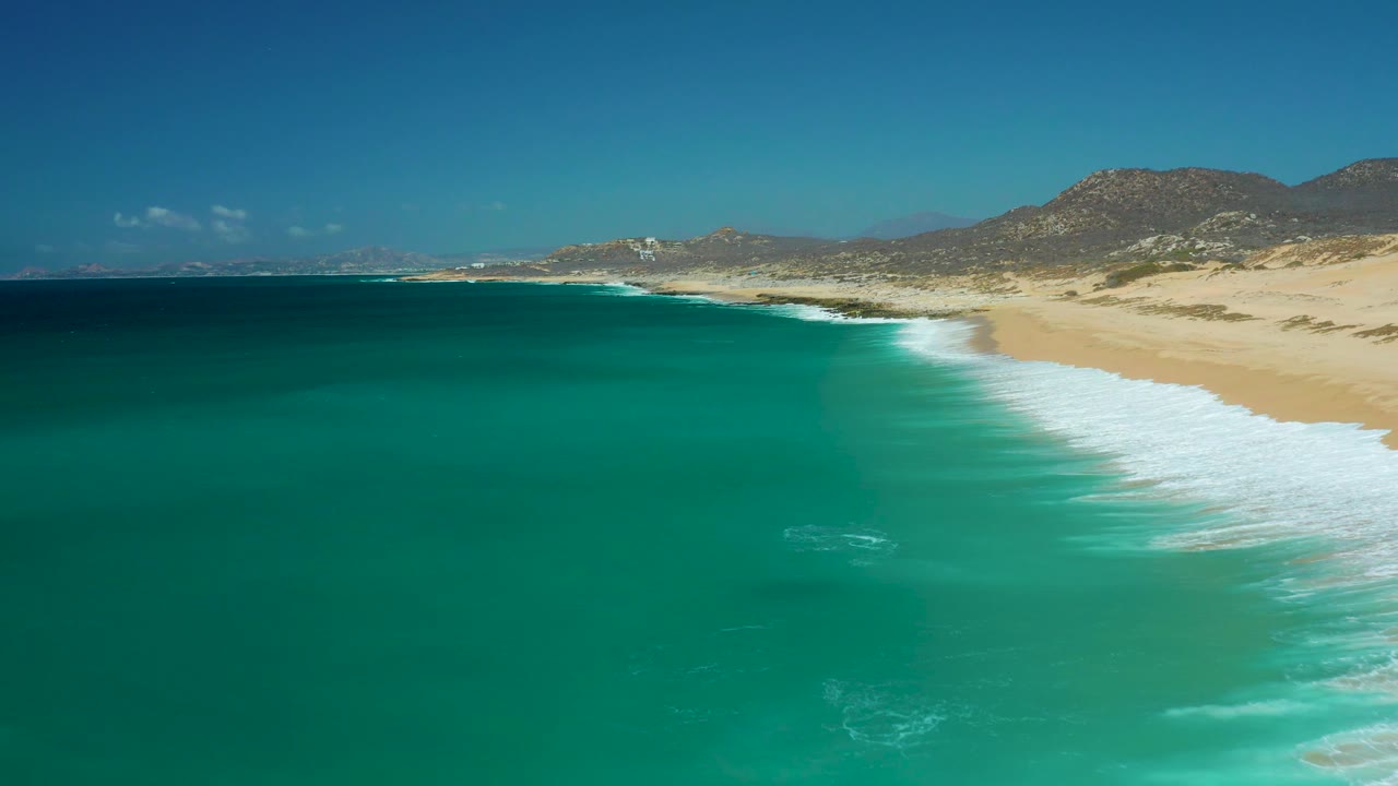 At La Laguna Beach Los Cabos, the aerial view holds turquoise water in the foreground and a pale sand beach with white foam on the right, backed by dunes and mountains, with no visible people.