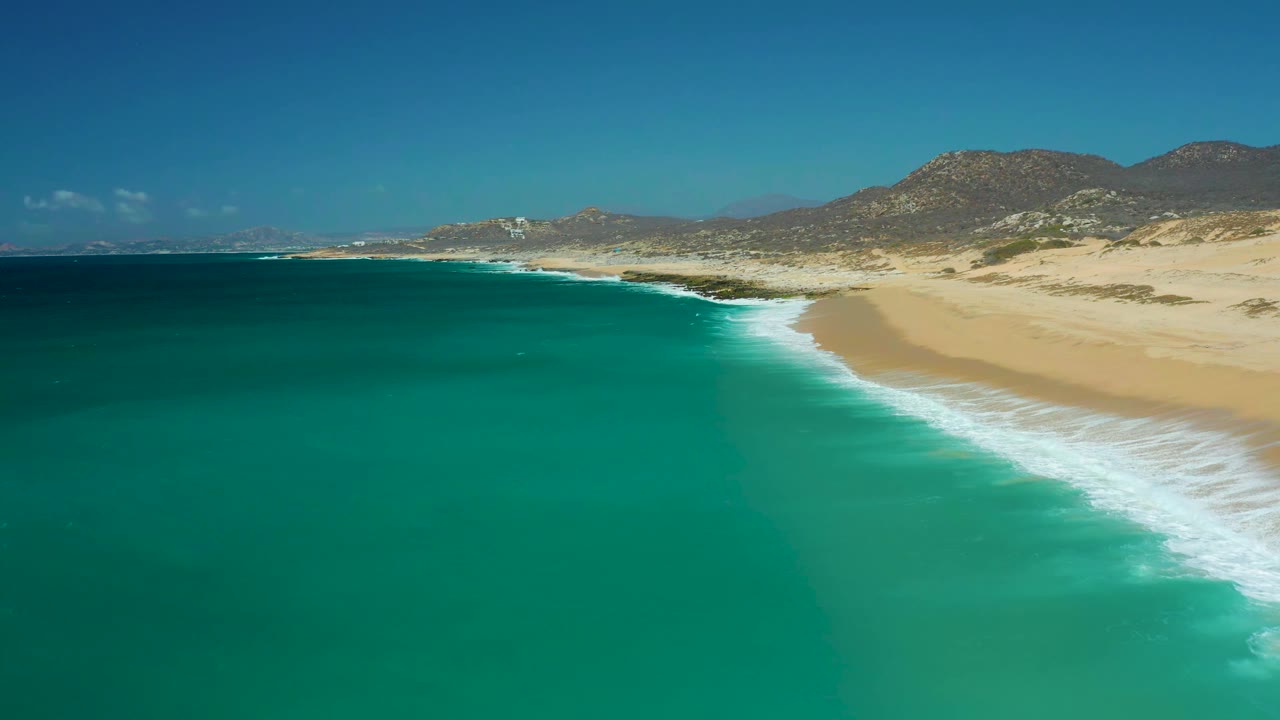 Aerial frame at La Laguna Beach in Los Cabos showing a diagonal band of white foam between deep turquoise water and open sand, with low mountains beyond and no visible people.